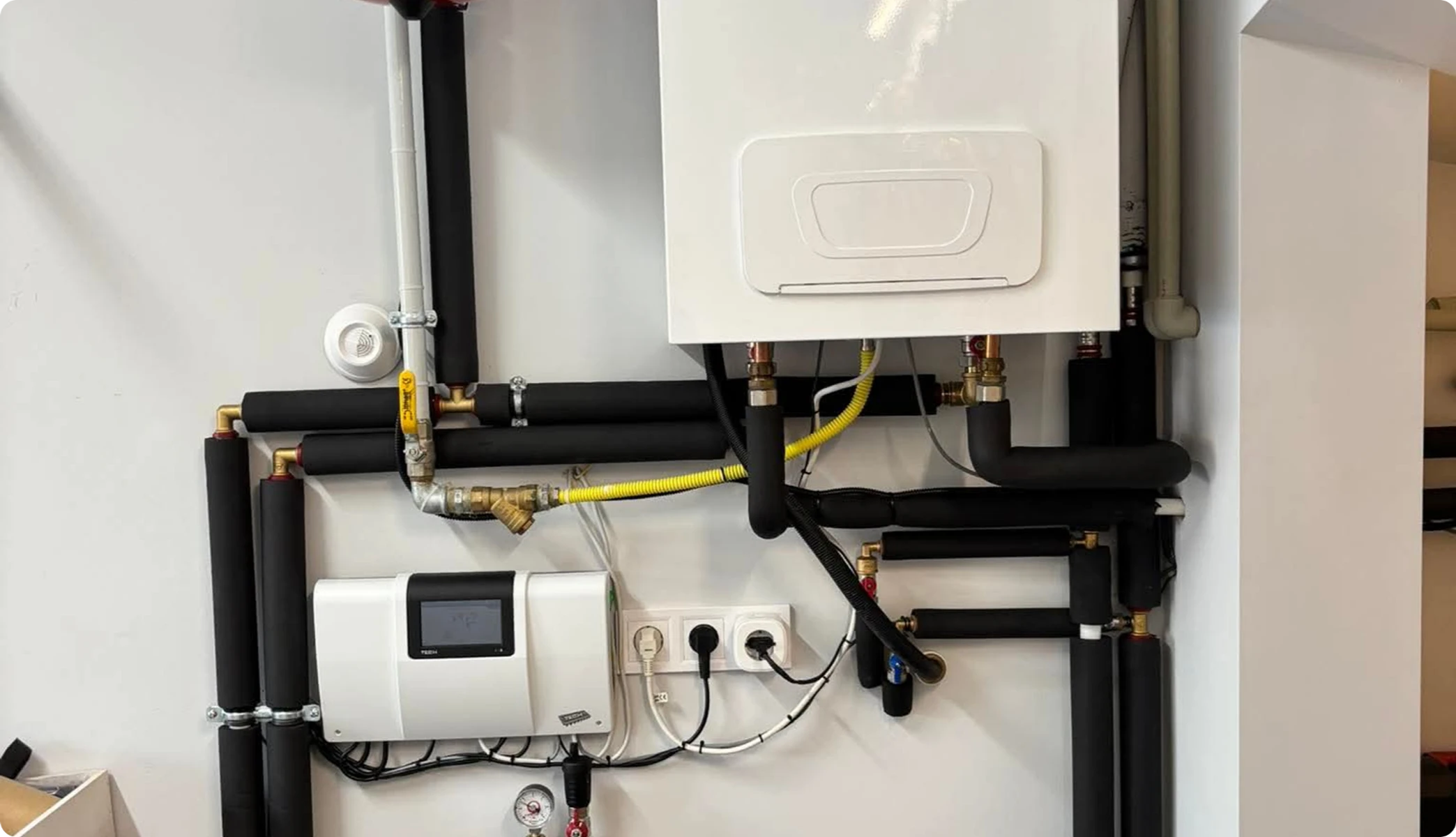 Biała ścienna jednostka kotłowa z izolowanymi czarnymi rurami i sterownikiem w systemie instalacji grzewczej